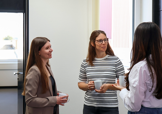 Drei Frauen stehen in einem Raum, zwei halten weiße Tassen, eine trägt ein gestreiftes Shirt, eine andere ein helles Hemd, die dritte einen Blazer.