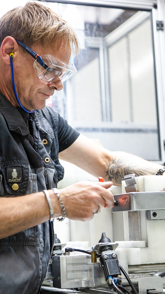 Handwerker mit Tätowierung an der rechten Unterarmaußenfläche bedient eine Maschine, trägt dunkelblaue Arbeitskleidung mit Mascot-Logo und Armband.