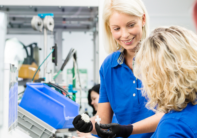 Zwei Personen in blauen Poloshirts und schwarzen Handschuhen halten weiße Kunststoffteile in einer Werkstatt oder Produktionsumgebung.