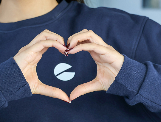 Hände formen ein Herz vor dunkelblauem Pullover mit weißem, kreisförmigem Logo.