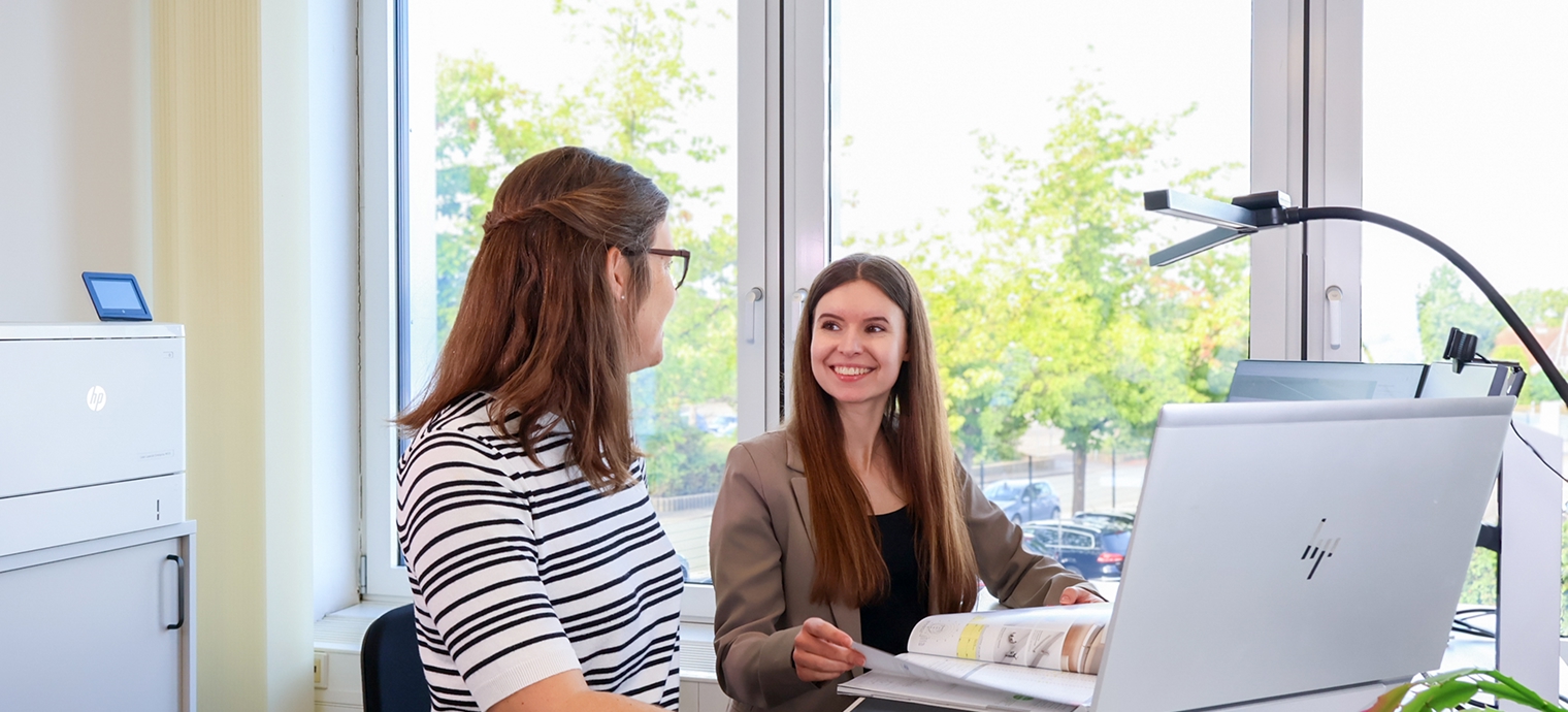 Zwei Frauen sitzen an einem Schreibtisch mit geöffnetem Ordner und Computer vor einem Fenster mit Blick auf Bäume und geparkte Autos