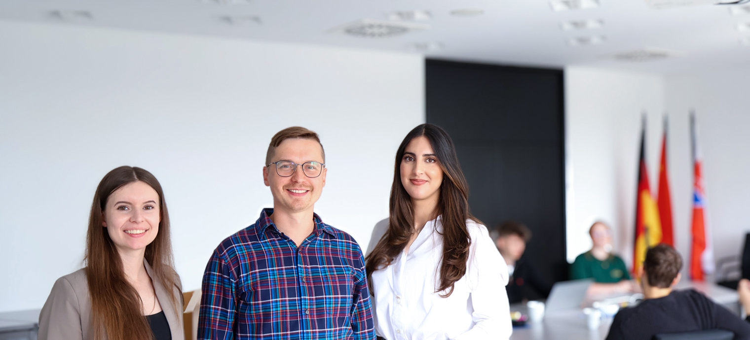 Drei berufserfahrene Mitarbeiterinnen und Mitarbeiter stehen nebeneinander in einem modernen Büro mit weiteren Personen im Hintergrund