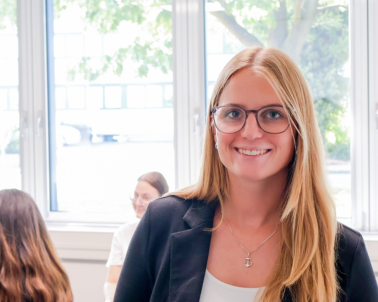 Junge Frau mit langem blonden Haar, schwarzem Blazer und Anker-Halskette vor großen Fenstern in einem hellen Raum mit weiteren Personen im Hintergrund.
