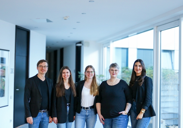 Fünf Personen stehen in einem hellen Flur nebeneinander und tragen lässige Kleidung mit dunklen Oberteilen und Jeans