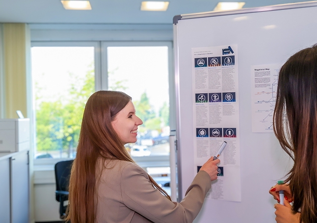 Zwei Frauen stehen vor einem Whiteboard mit ausgedruckten Informationsblättern, eine zeigt mit einem Stift darauf